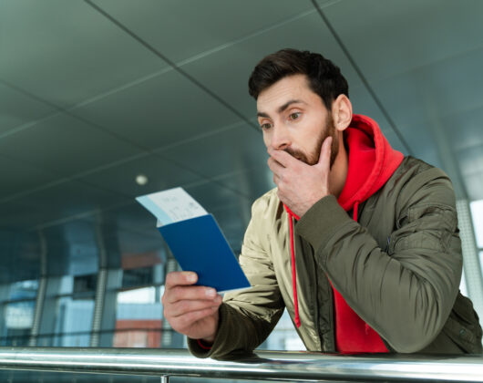 Waist up portrait of shocked man standing at the airport and looking at his passport with surprised expression while waiting for departure. Technologies and people concept